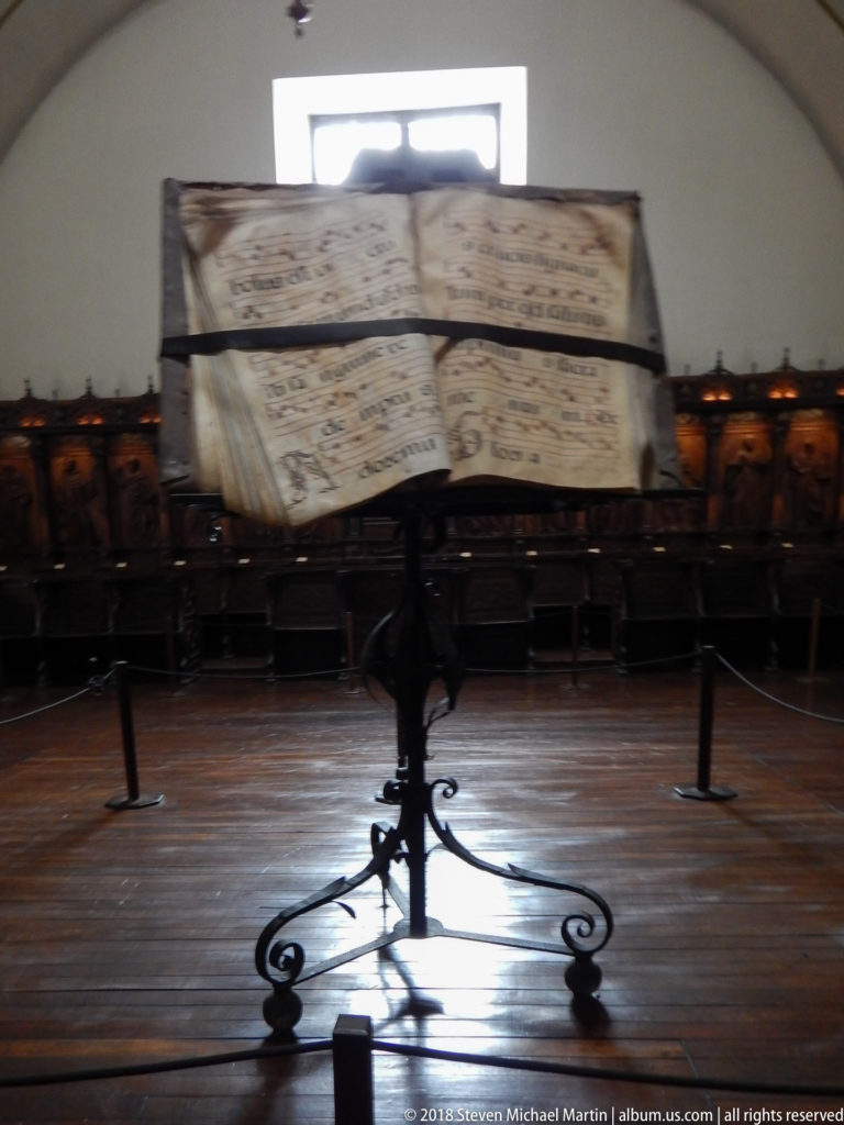 Large open choir book with musical notation on an ornate iron stand in a wood-paneled room.