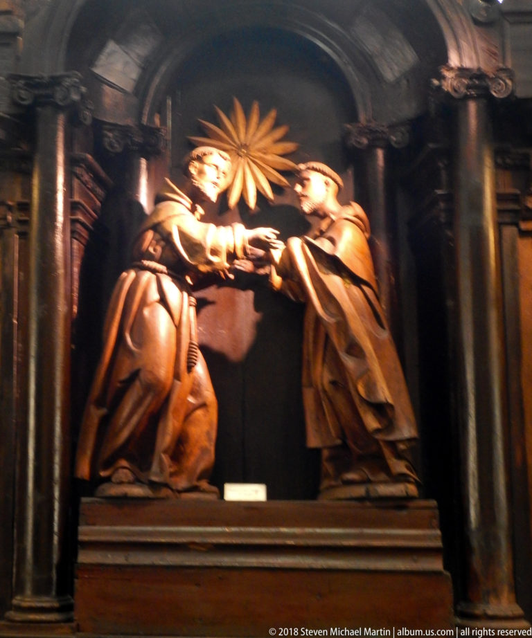 Close view of carved wooden relief showing two kneeling friars facing each other beneath a radiant emblem.