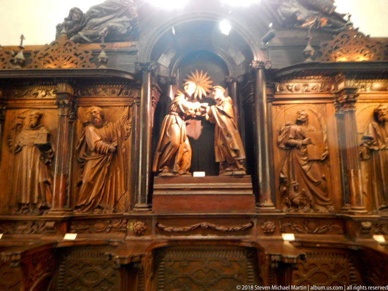 Dark carved wooden devotional structure with a haloed central figure in a niche and relief panels of saints on both sides.