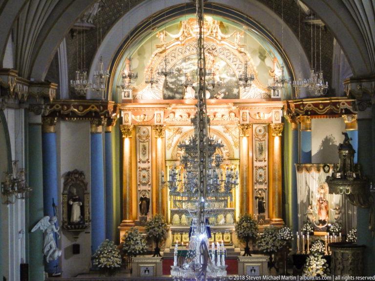 Large chandelier centered before Santo Domingo’s illuminated high altar with gilded columns, flowers, and side devotional figures.