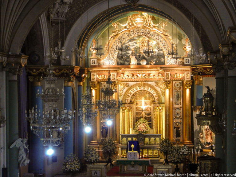 Illuminated high altar and sanctuary with gilded ornament, columns, chandeliers, side statues, and floral arrangements.