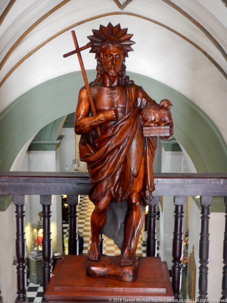 Carved wooden Saint Christopher carrying the Christ Child and holding a staff, displayed against a pale interior arch.