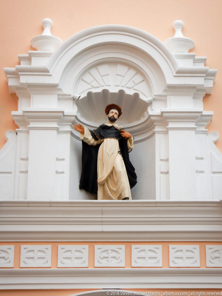 Polychrome statue of Saint Dominic in black-and-white Dominican habit, standing with arms extended in a white façade niche.