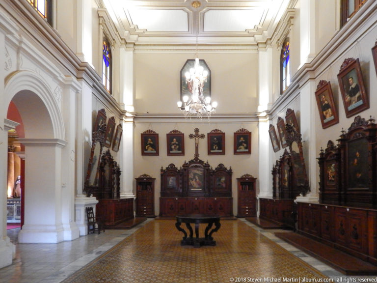 Formal white-walled monastery room with carved wooden furnishings, devotional paintings, chandelier, and central table.