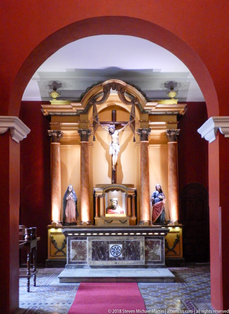 Side chapel seen through an arch, with crucifix, flanking saint statues, central shrine, and marble-fronted altar.