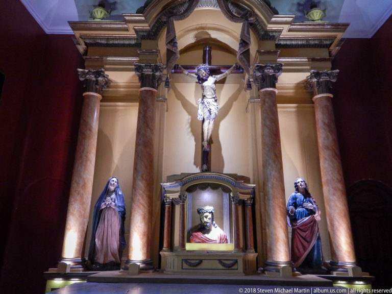 Chapel altar with large crucifix, central bust of Christ, flanking saint statues, columns, and dark red wall.
