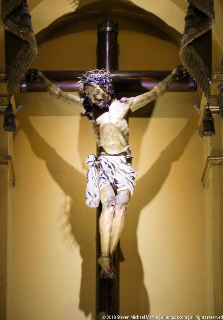 Polychrome crucifix of Christ against an ochre wall, with a strong cross-shaped shadow cast behind the figure.