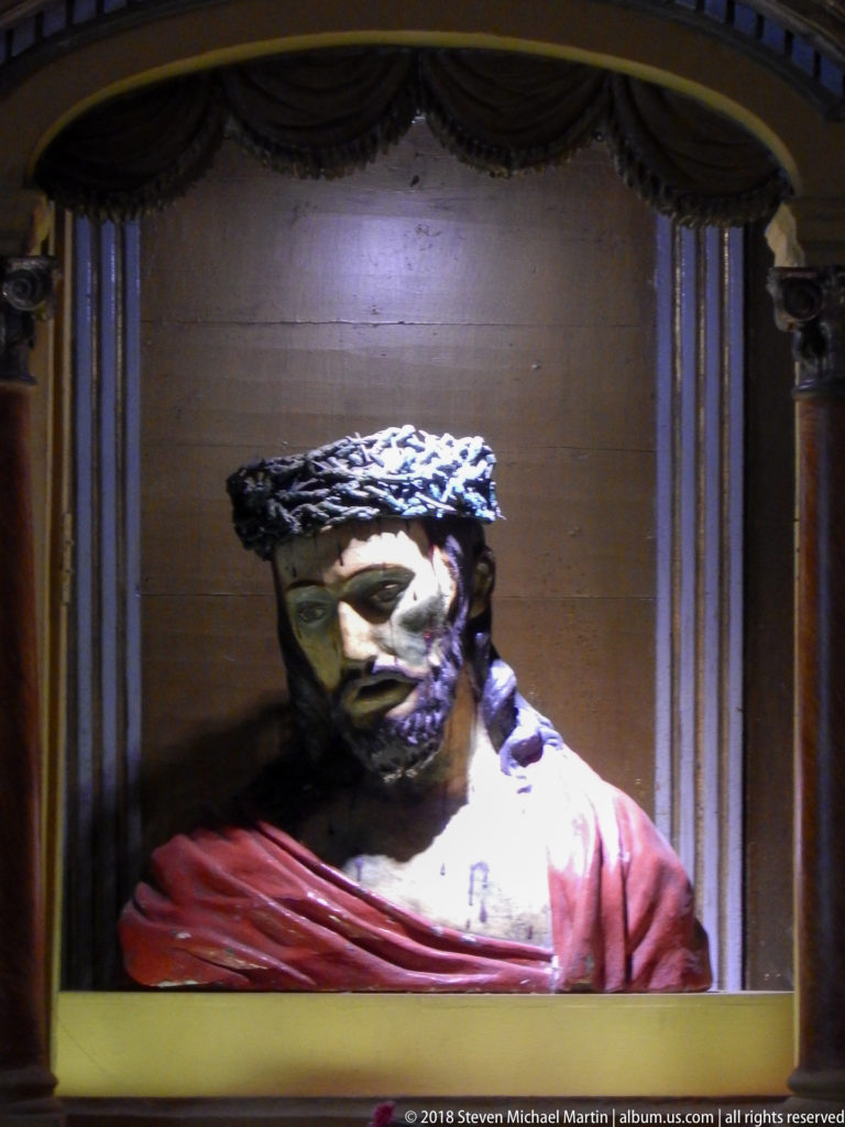 Polychrome bust of Christ with crown of thorns, painted wounds, and red cloak in an architectural niche.