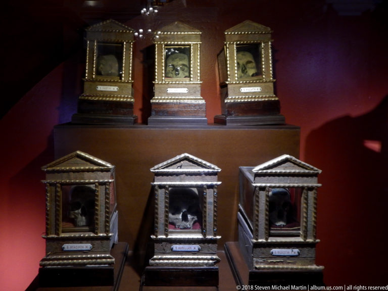 Six small architectural reliquary cases with pediments and gilded trim displayed against a dark red wall.