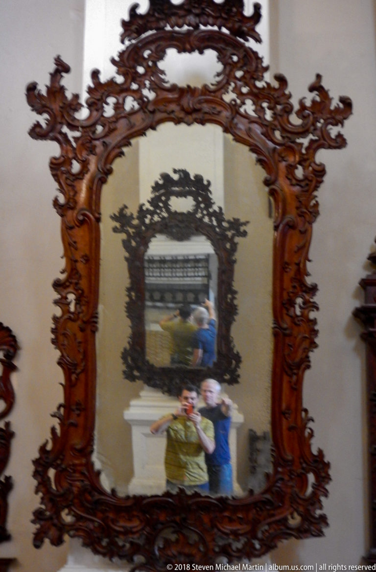 Large carved wooden mirror with scrolling ornament reflecting visitors and another ornate mirror in the Santo Domingo complex.