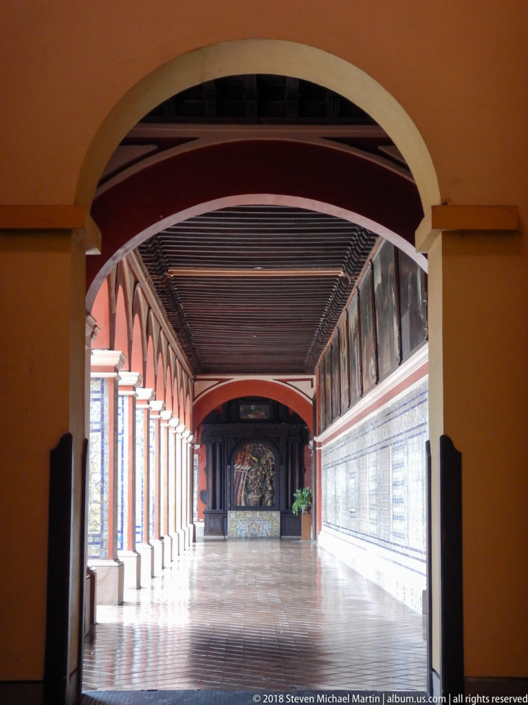 Arcaded cloister corridor with tiled wall, religious paintings, timber ceiling, and polished floor at Santo Domingo in Lima.