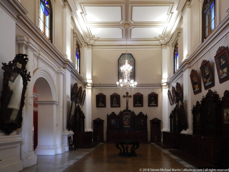 High-ceilinged convent room with carved dark furniture, framed portraits, chandeliers, stained-glass windows, and a central table.