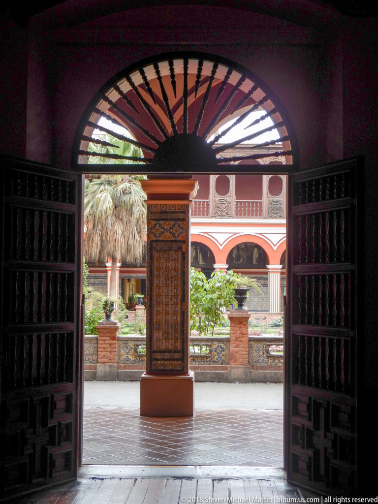 Dark wooden doorway with fan-shaped transom framing Santo Domingo’s bright cloister garden and red arcades beyond.