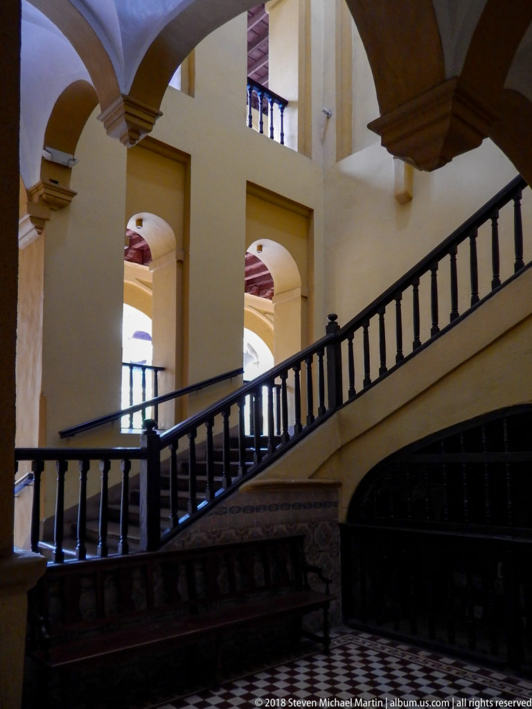 Dark wooden staircase and balustrade crossing a pale yellow convent interior with white arches, gallery openings, and patterned floor.