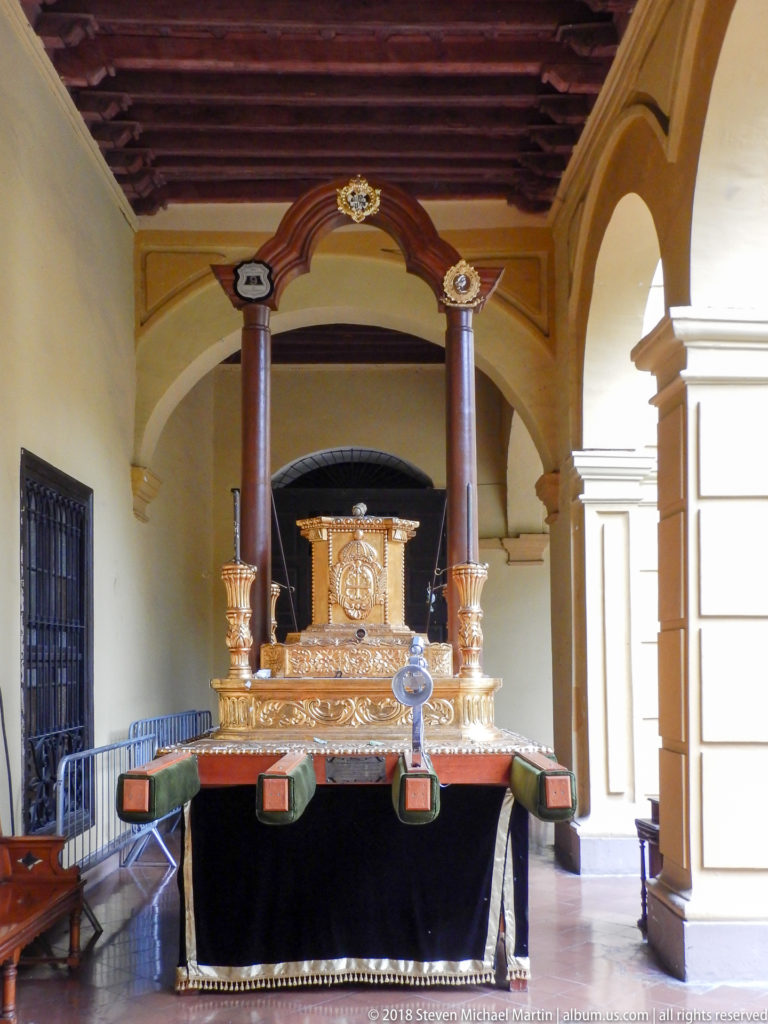 Gilded portable devotional structure with slender columns and projecting carrying beams displayed beneath a convent arcade.