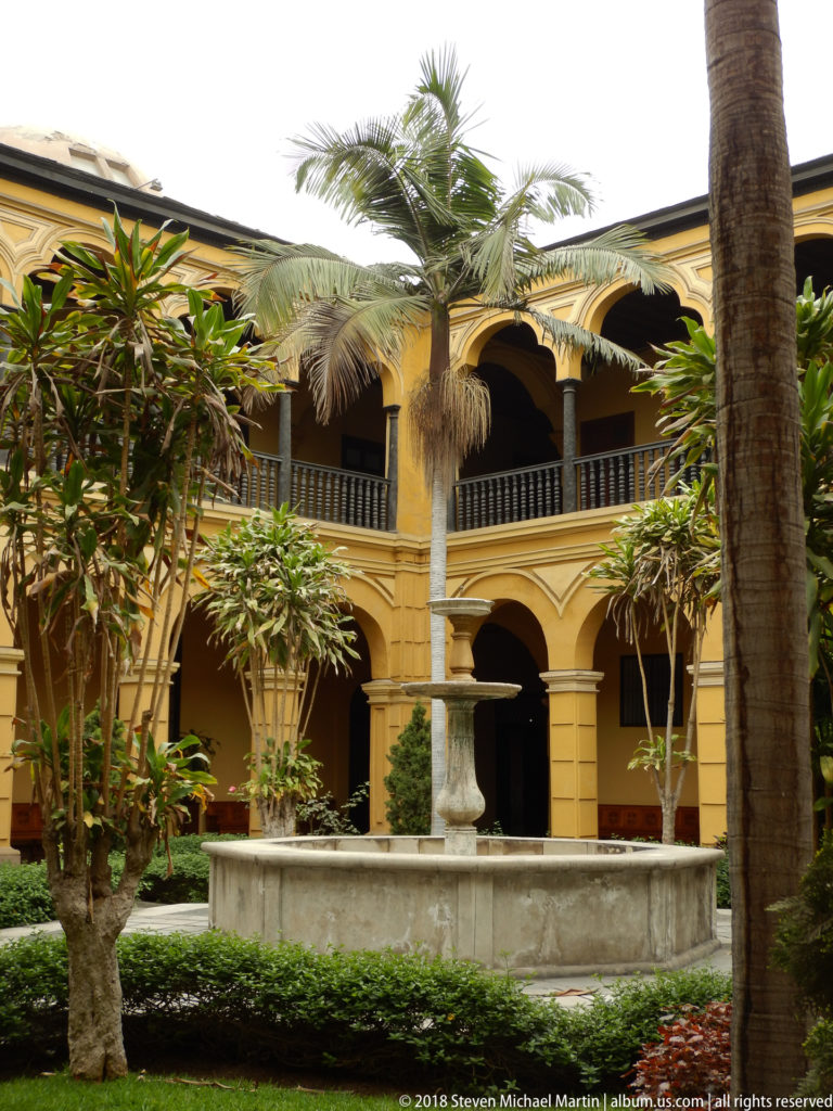 Two-story ochre cloister around a planted courtyard with palms, hedges, and a broad central stone fountain basin.