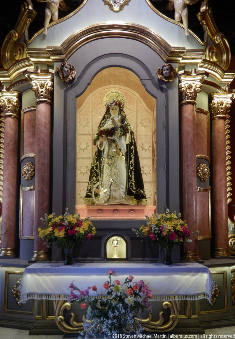 Close view of a richly dressed Marian statue in an arched altar niche, framed by red columns and flowers.