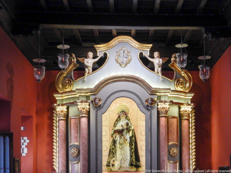 Ornate Marian altar with dressed female sacred figure, paired red columns, gilded capitals, angel figures, and hanging glass lamps.