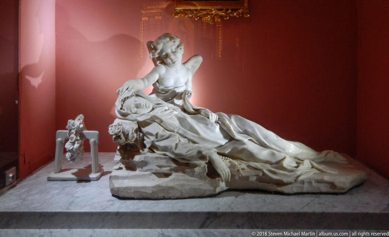 Full view of a white-stone funerary monument with recumbent figure and winged angel, accompanied by smaller sculptures against red walls.