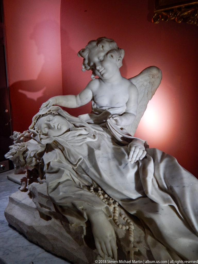 White-stone funerary sculpture of a recumbent female figure with a winged angel rising beside her against a dark red wall.