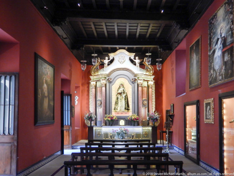 Small red-walled chapel with wooden benches leading to an ornate altar containing a richly dressed statue of the Virgin.