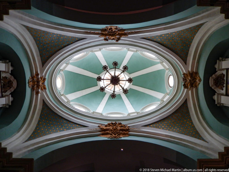 Oval turquoise-and-white dome with radial ribs, small oval windows, central chandelier, and gilded ornament around the surrounding arches.