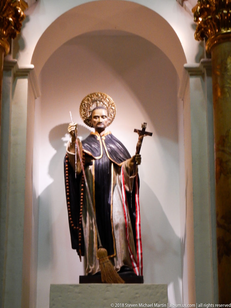Statue of Saint Martin de Porres in Dominican habit with halo, broom, and crucifix, displayed in a shallow arched niche.