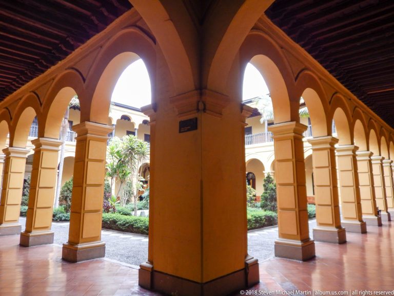 Ochre cloister arcade with repeated semicircular arches opening onto a planted courtyard with palms and shrubs.