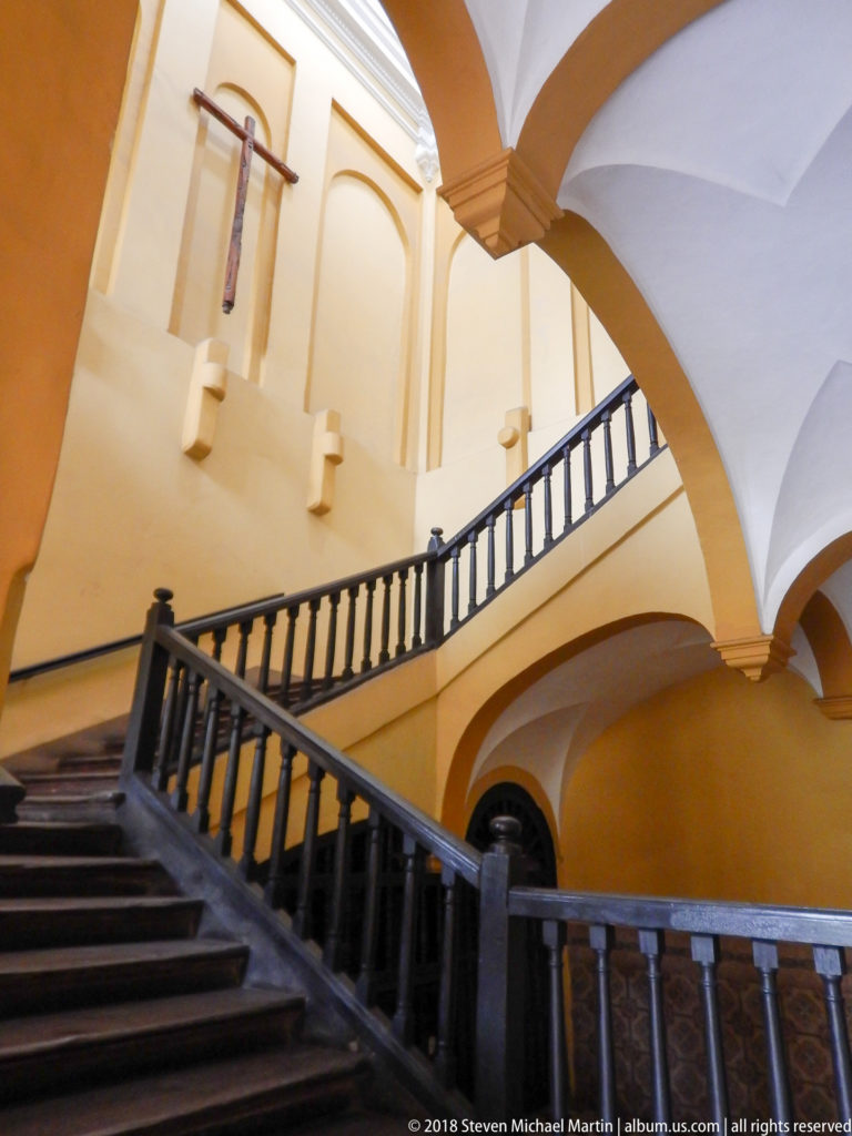 Broad convent staircase with dark wooden balustrade, pale yellow walls, arches, white vaults, and high windows.