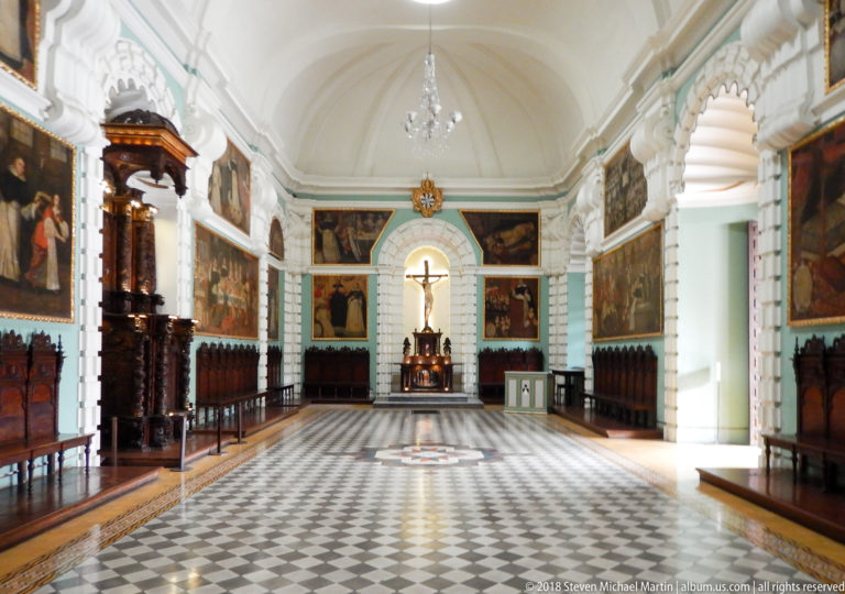 Long convent hall with checkerboard floor, central star medallion, carved wooden stalls, religious paintings, and a crucifix at the far end.