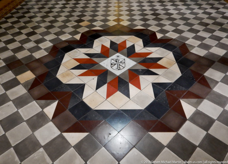 Eight-pointed red, cream, gray, and black floor star set within a checkerboard stone pavement, with a small black-and-white emblem at center.