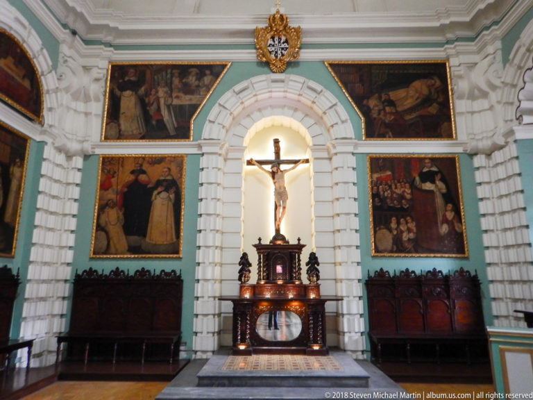 Crucifix in a tall white arched niche, centered above a dark wooden pedestal and surrounded by framed religious paintings and carved seating.