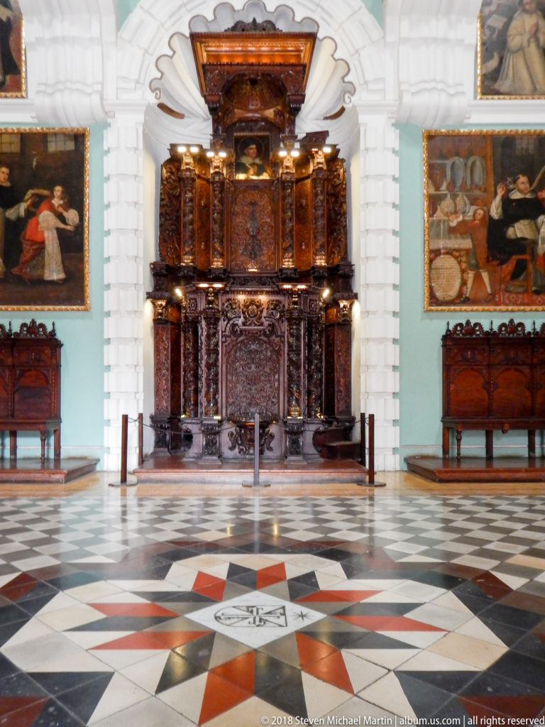 Large dark carved wooden furnishing with columns and panels, flanked by religious paintings and benches on a geometric stone floor.