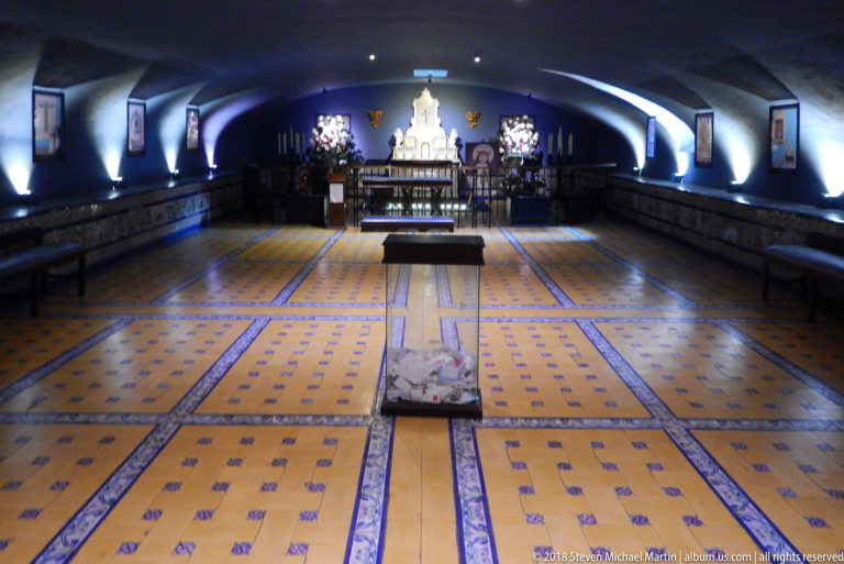 Long vaulted crypt with patterned floor, rows of burial markers, side benches, a central display opening, and altar at the far end.