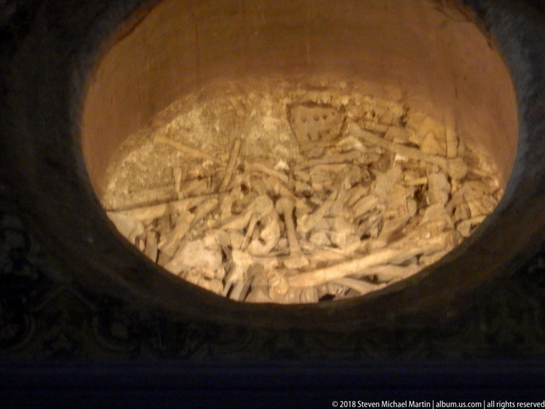 Circular crypt opening densely filled with disarticulated long bones and smaller skeletal fragments under warm light.