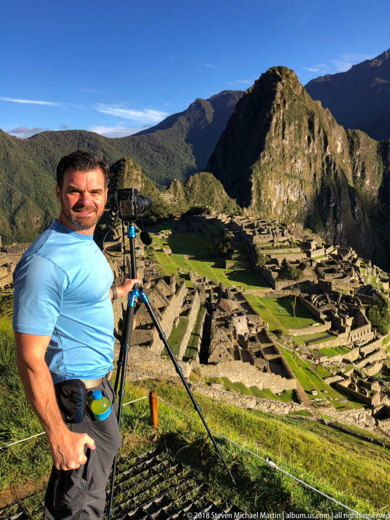 Visitor holding a tripod beside a camera with Machu Picchu and Huayna Picchu filling the background.