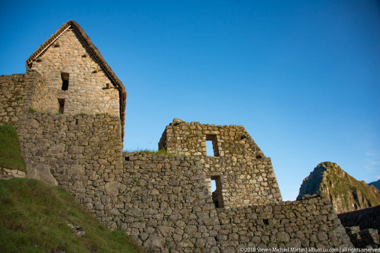 Gabled thatched stone building beside a roofless masonry structure with rectangular openings at Machu Picchu.