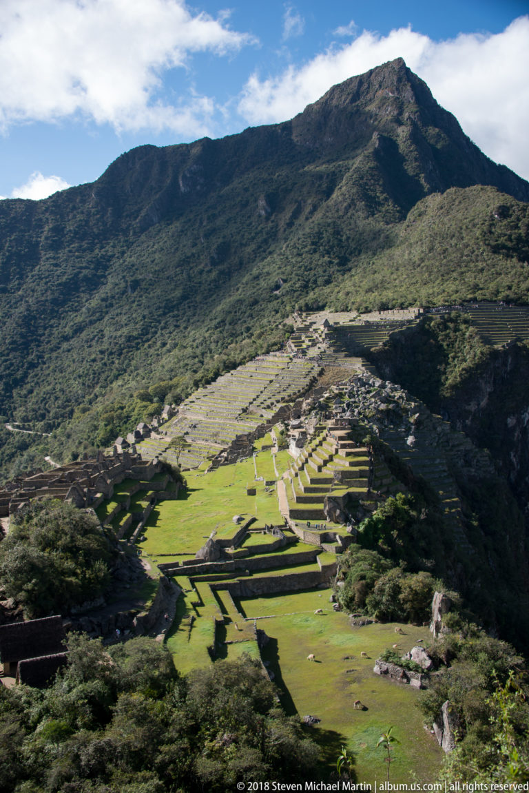 Machu Picchu’s sunlit terraces and buildings lie beneath a broad forested Andean mountain ridge.