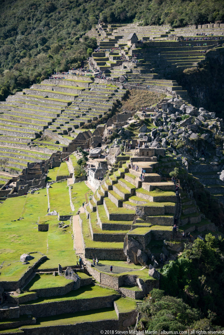Elevated view of Machu Picchu with long terraces, open lawns, paths, and dense stone buildings.