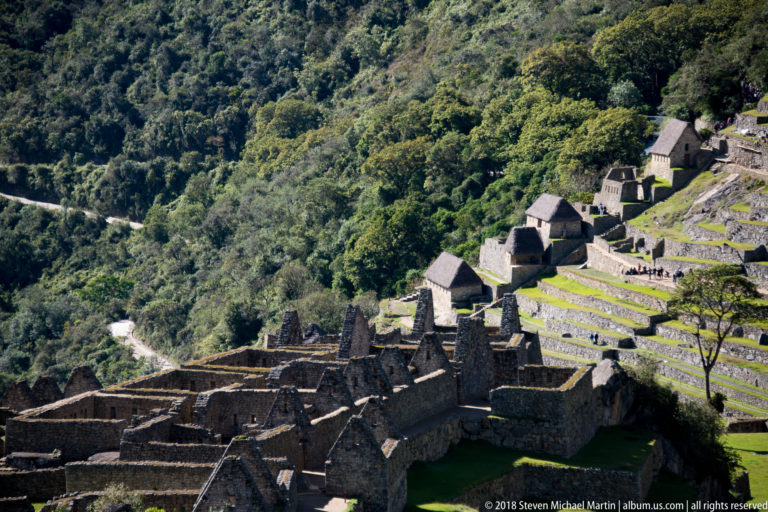 Roofless stone compounds below long green terraces and a few thatched roofs at Machu Picchu.