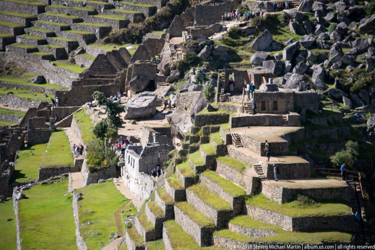 High-angle view of Machu Picchu stone rooms, passages, terraces, boulders, and visitors clustered on the slope.