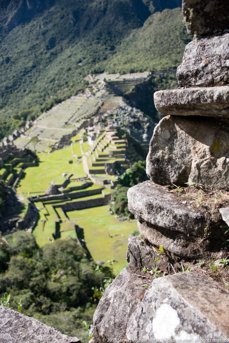 Foreground stone masonry on Huayna Picchu overlooks Machu Picchu’s terraces, lawns, and buildings far below.