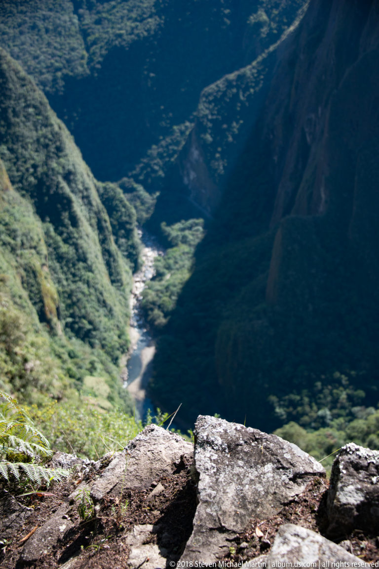 Steep view from a rocky mountain edge down to the river winding through the forested Urubamba valley.