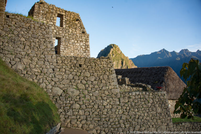 Layered stone retaining walls and a thatched roof at Machu Picchu with Huayna Picchu beyond.