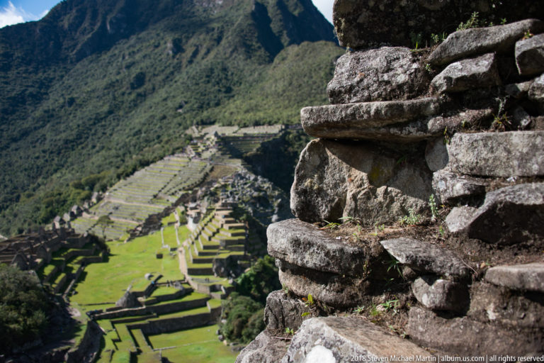 Close stone masonry wall on Huayna Picchu with Machu Picchu’s terraces and buildings blurred far below.
