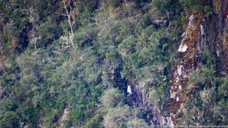 Distant visitor on a narrow cliffside path amid dense vegetation and exposed rock on Huayna Picchu.