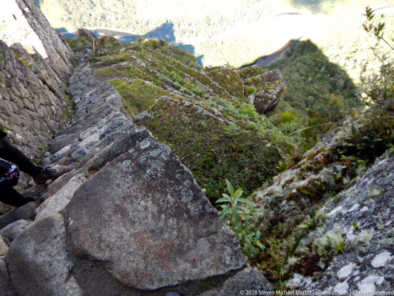 Large boulders and rough stone edging crowd a narrow, vegetation-lined section of the Huayna Picchu trail.