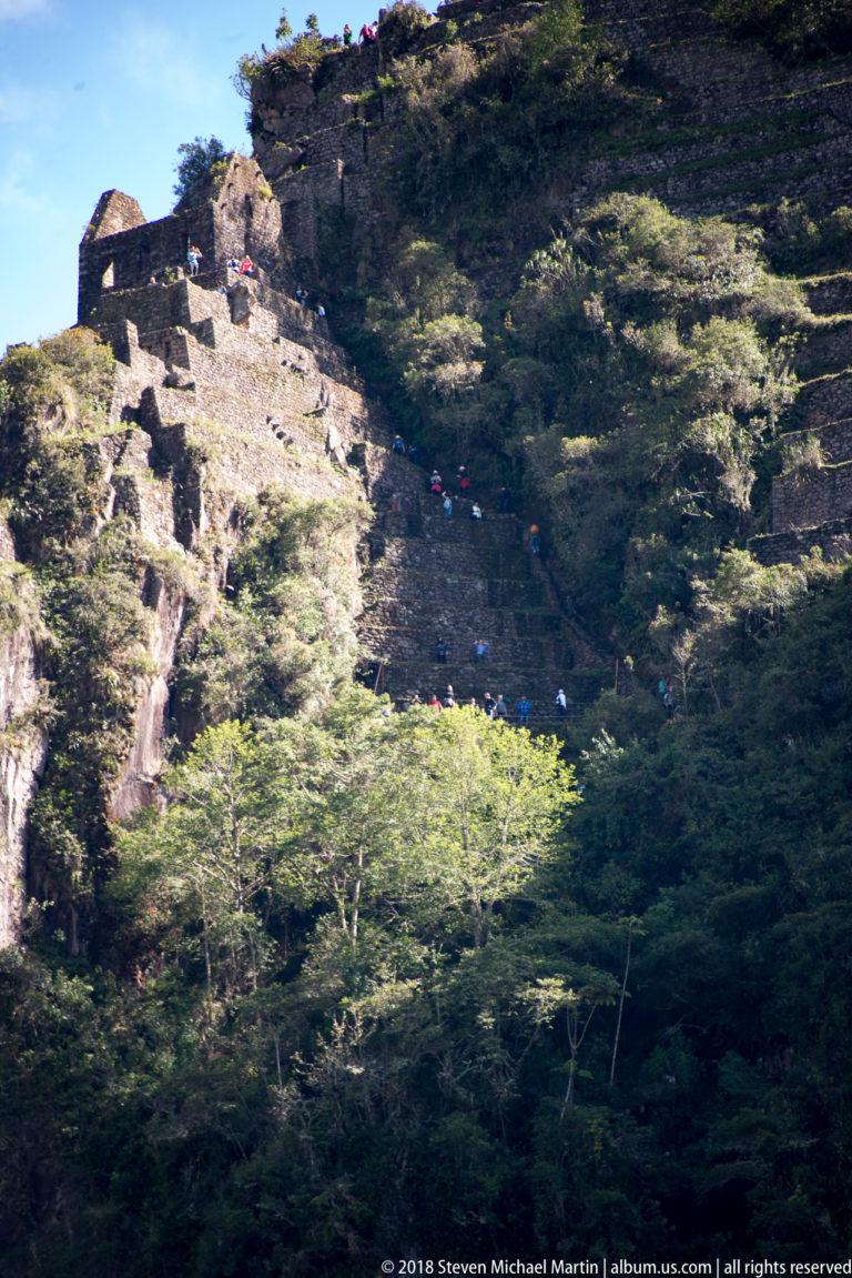 Oblique view of Huayna Picchu’s steep stone route with visitors beside retaining walls and dense vegetation.