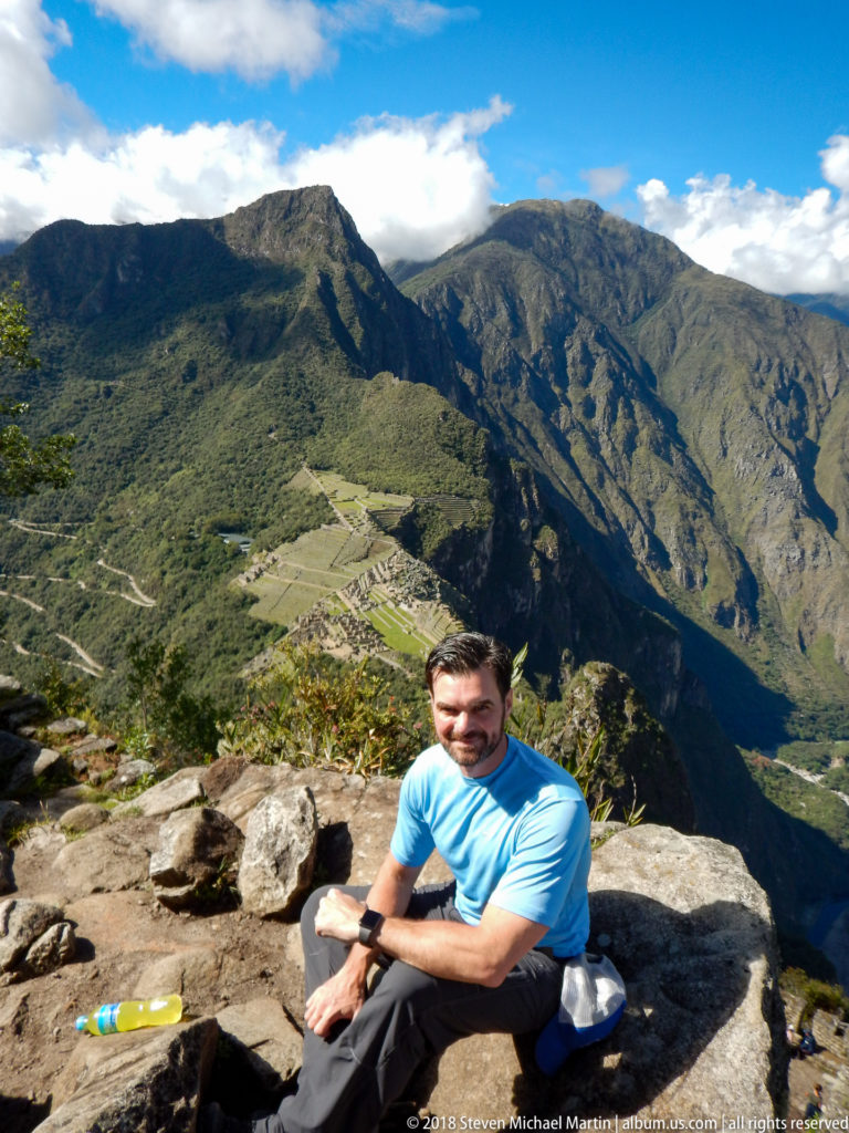 Seated visitor on a rocky Huayna Picchu ledge with Machu Picchu and green mountains visible far below.