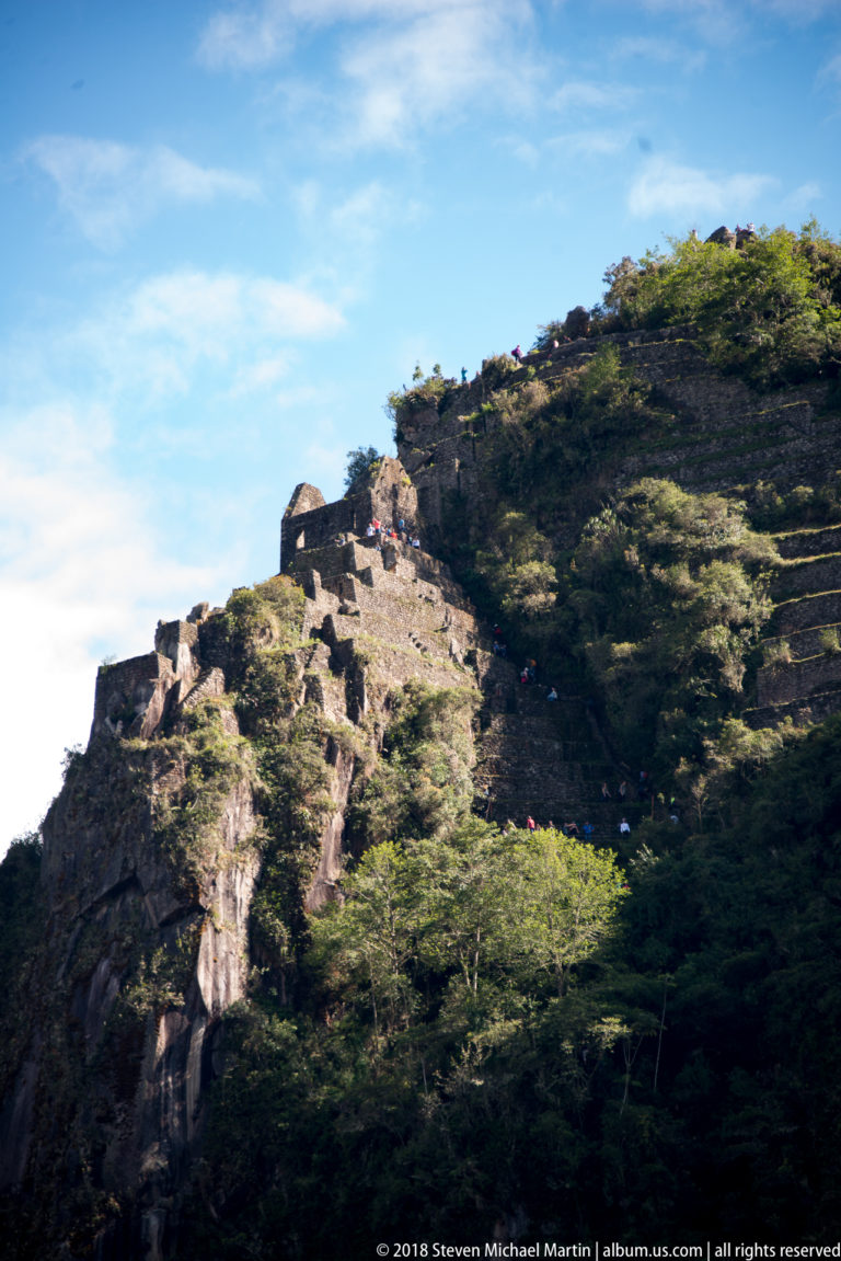 Upper Huayna Picchu slope with stone terraces, small structures, vegetation, and visitors along the ridge.